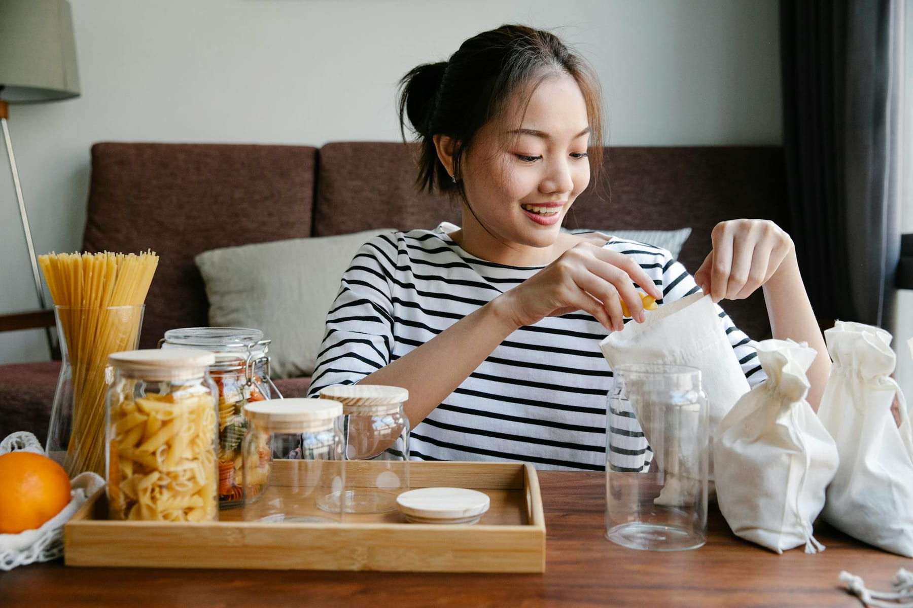 Smiling Asian woman opening environmentally friendly bags of pasta while sitting near glass jars with pasta on table at home on Earth Wise Budget