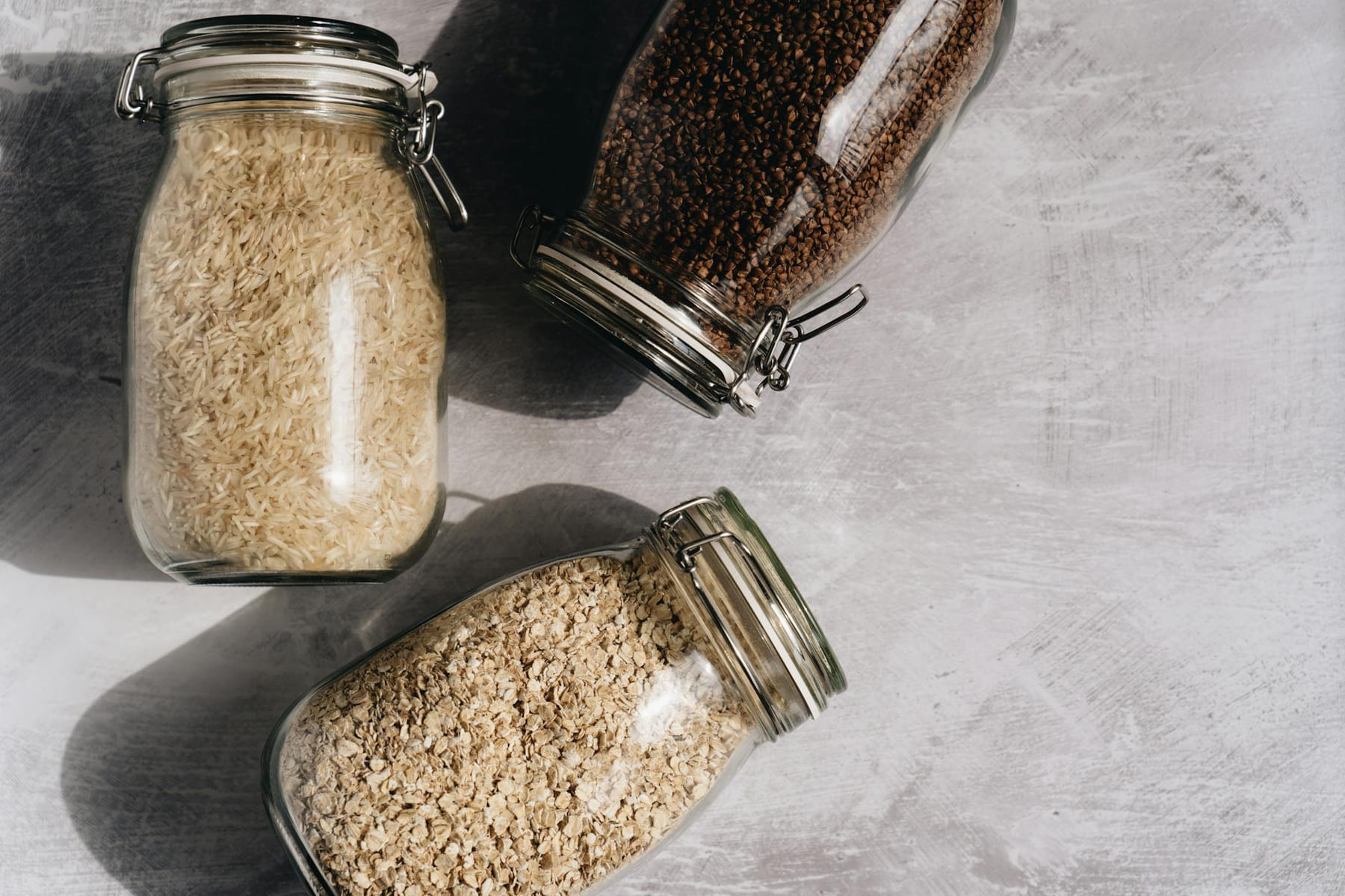 Top-down view of rice, oats, and buckwheat in glass jars on a textured surface. on Earth Wise Budget